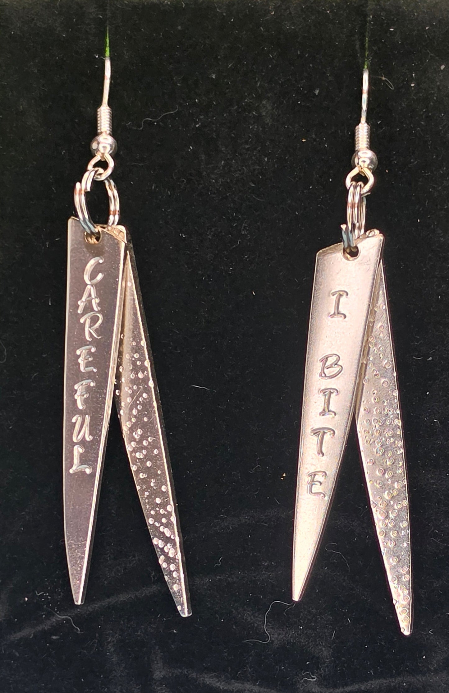 A pair of silver dangling earrings displayed on a black velvet jewelry stand. Each earring has a fishhook wire and a pair of pointed silver charms, one smooth and one textured, with engraved text reading 'CAREFUL' and 'I BITE'.