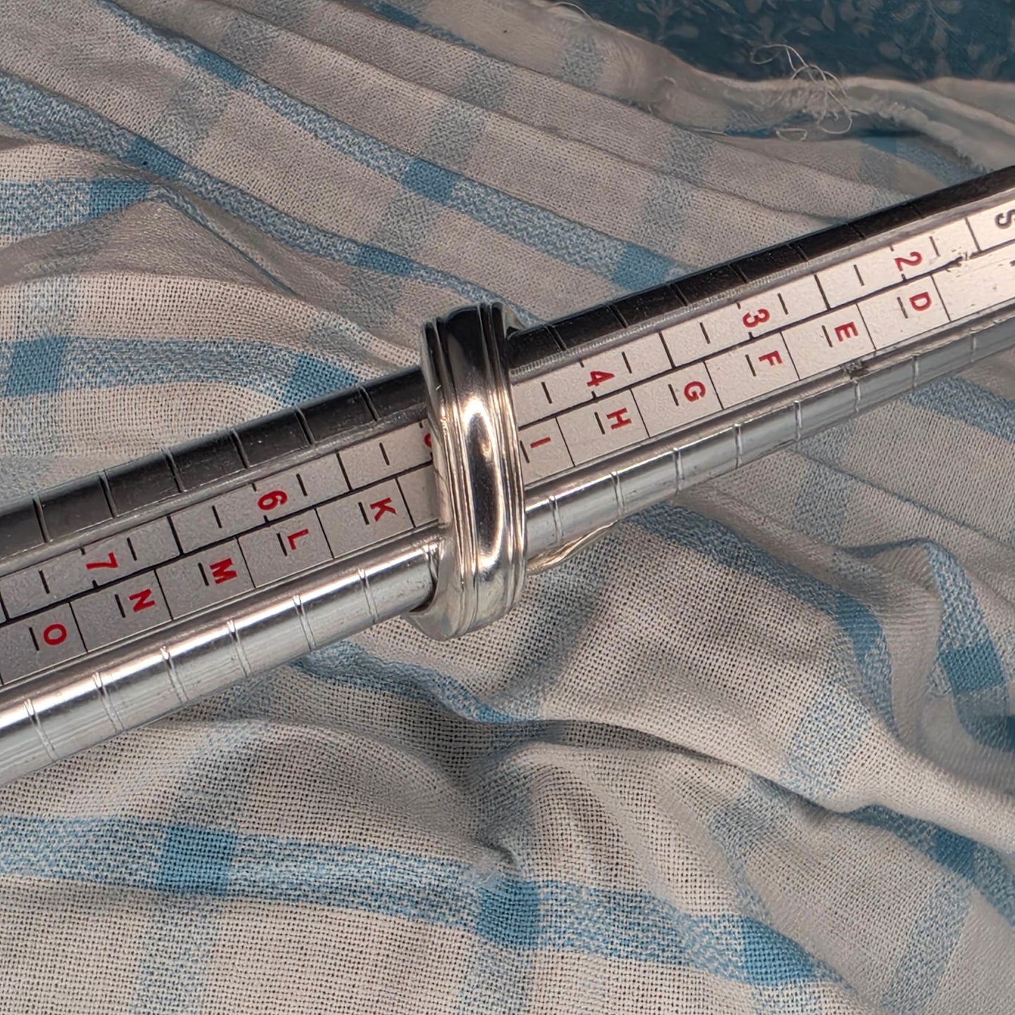 A silver-toned metal ring displayed on a sizing guide labeled with letters from 2D to NO, resting against a light blue and white striped fabric background.