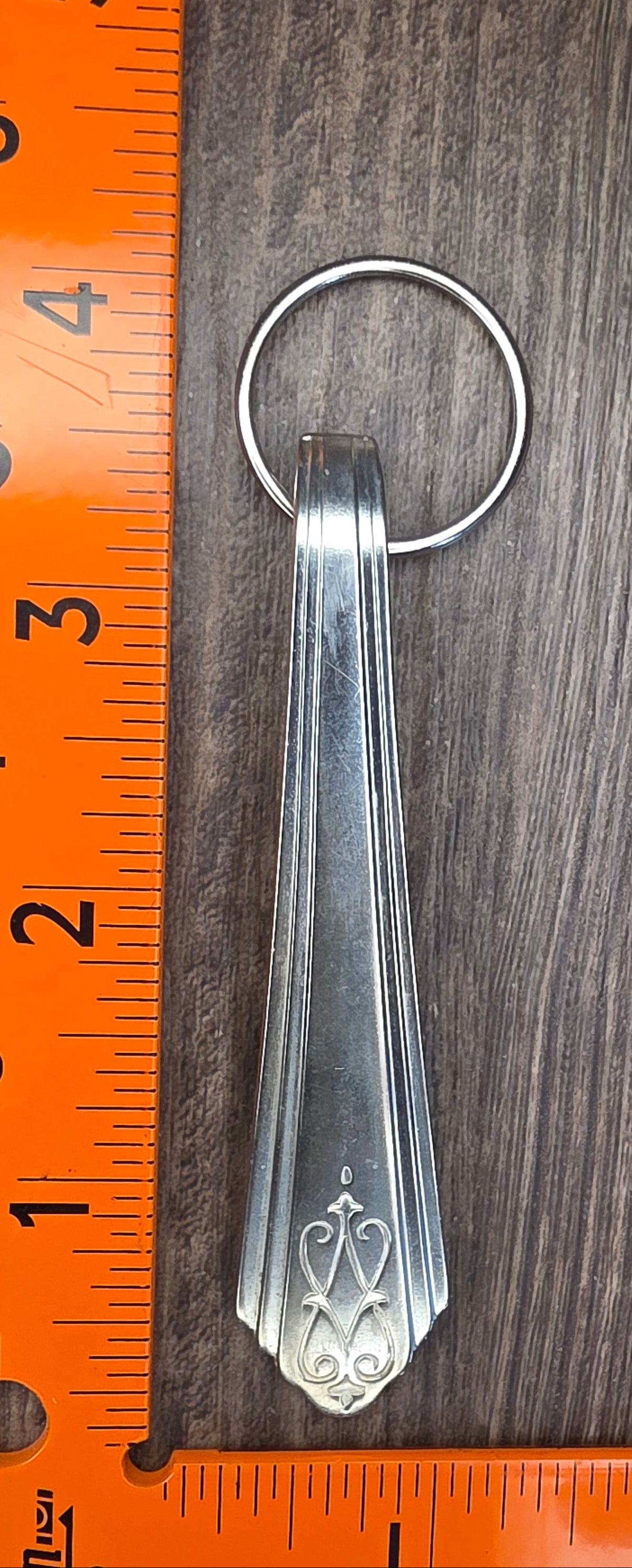 Silver keychain with decorative design on a wooden surface with an orange ruler for scale.