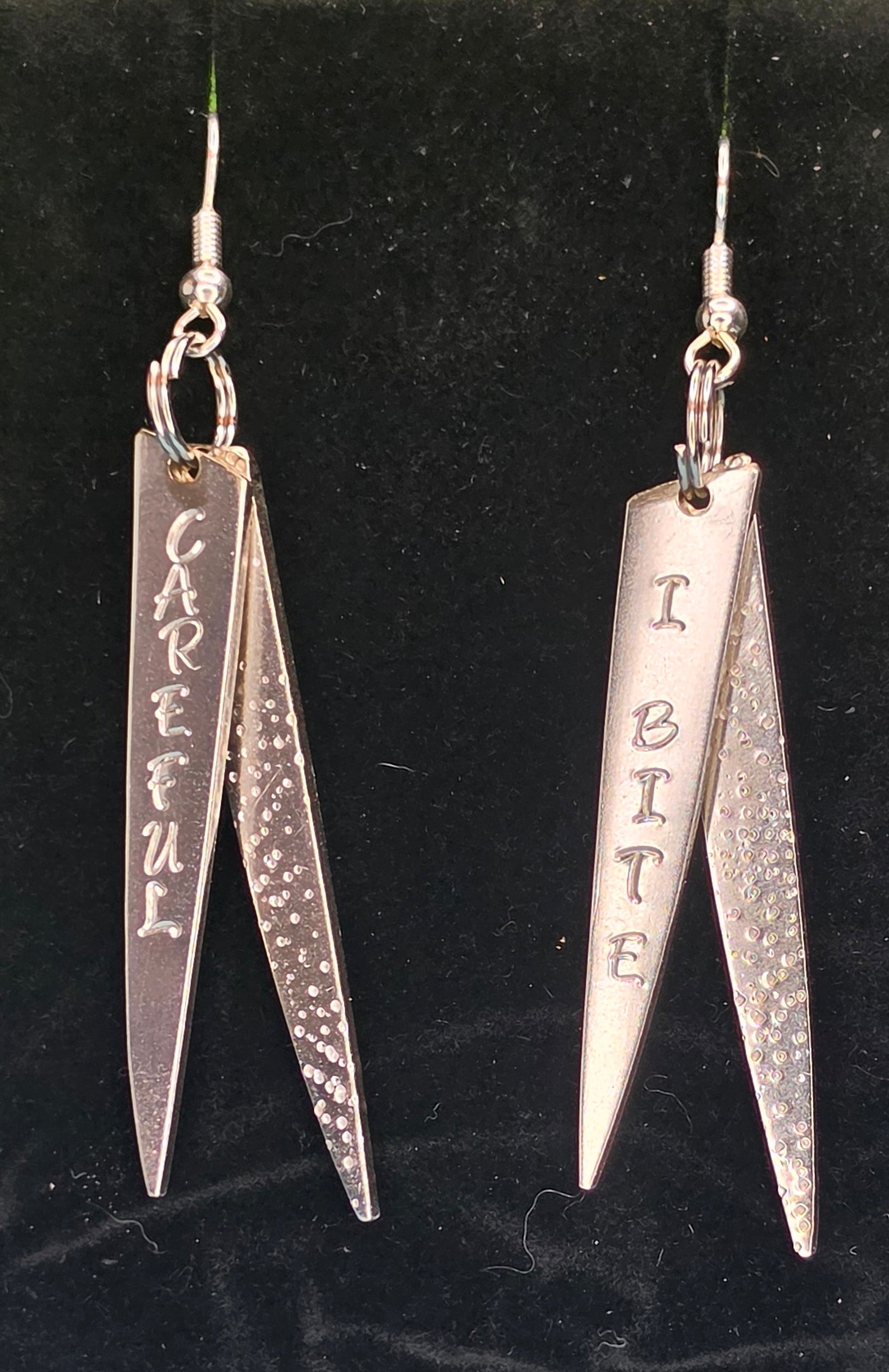 A pair of silver dangling earrings displayed on a black velvet jewelry stand. Each earring has a fishhook wire and a pair of pointed silver charms, one smooth and one textured, with engraved text reading 'CAREFUL' and 'I BITE'.