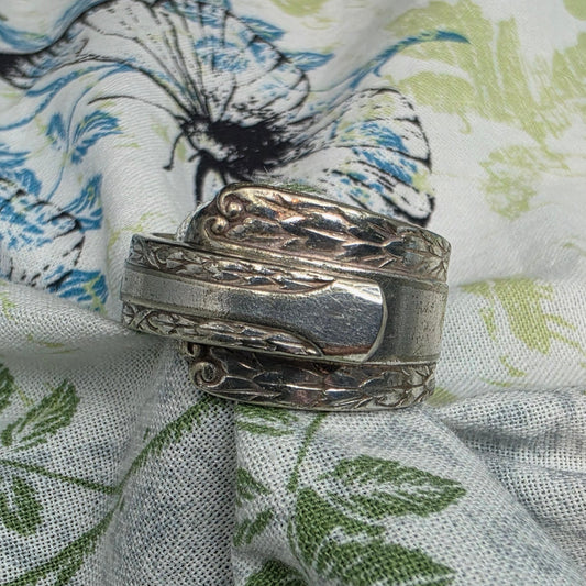 A wide silver-toned band ring with engraved floral and scroll details, displayed on a white fabric background with a blue, green, and gray floral print.