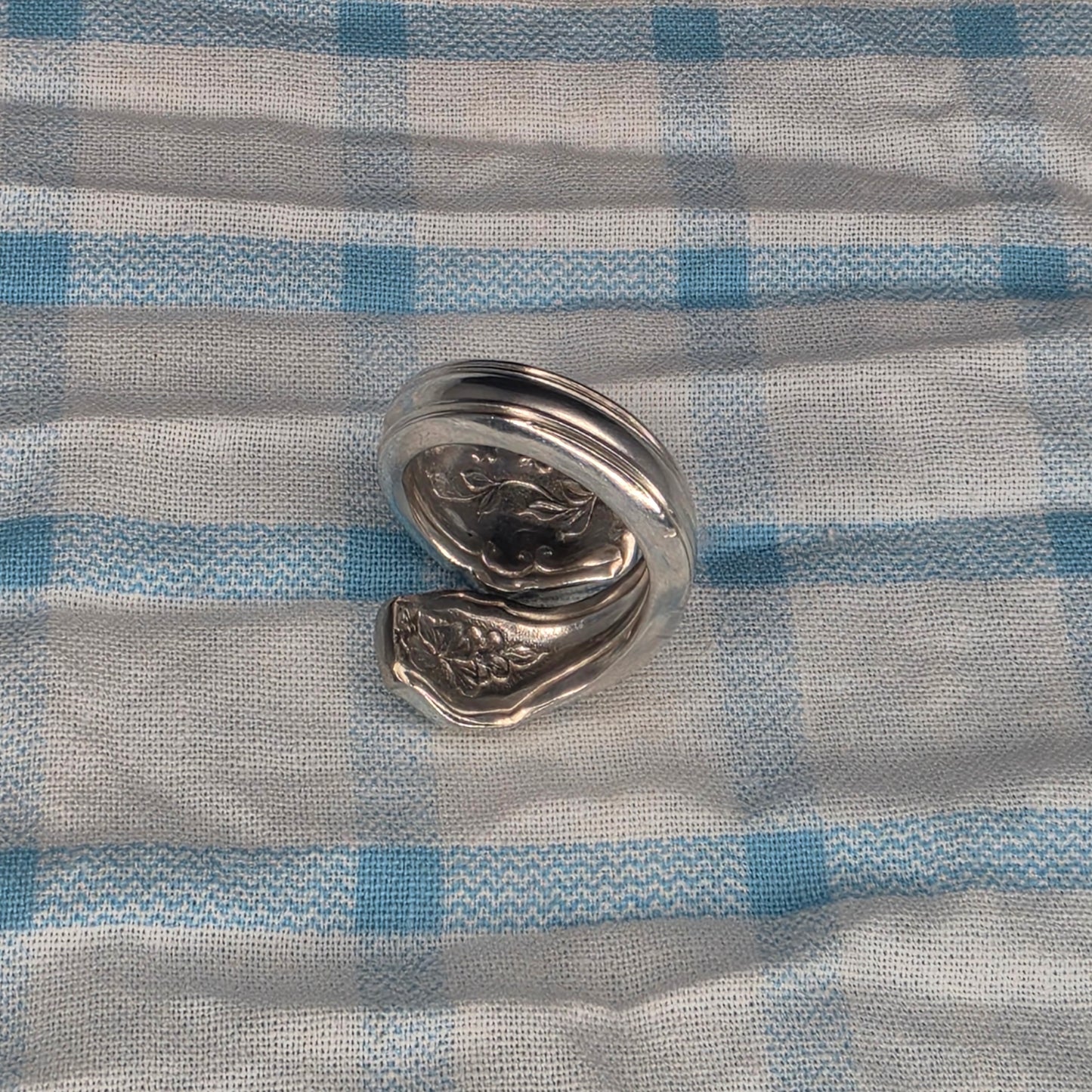 A polished silver metal ring with a textured, organic shape, displayed on a blue and white checkered fabric background.