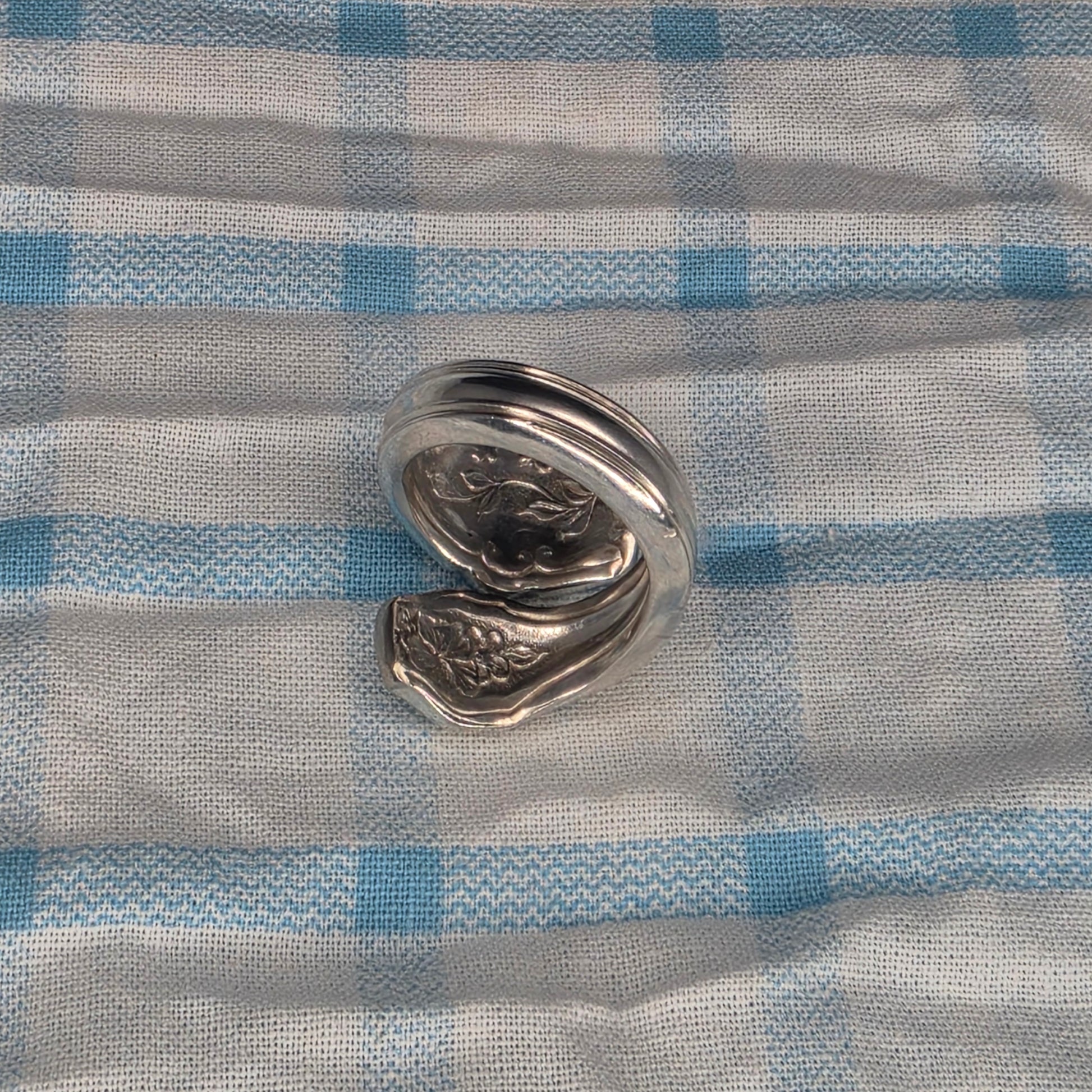 A polished silver metal ring with a textured, organic shape, displayed on a blue and white checkered fabric background.