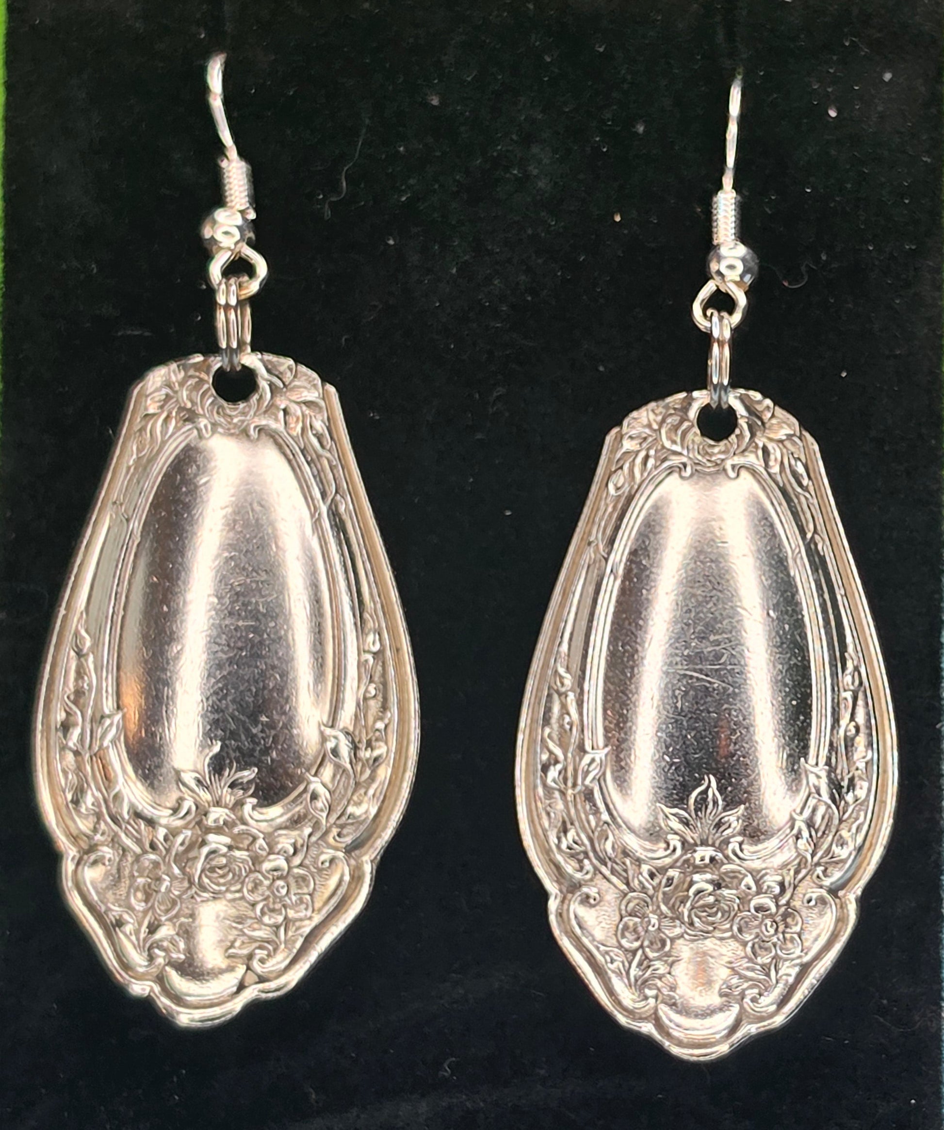 A pair of silver-toned drop earrings shaped like vintage ornate spoon bowls, displayed on a black velvet jewelry stand against a green and blue patterned background.