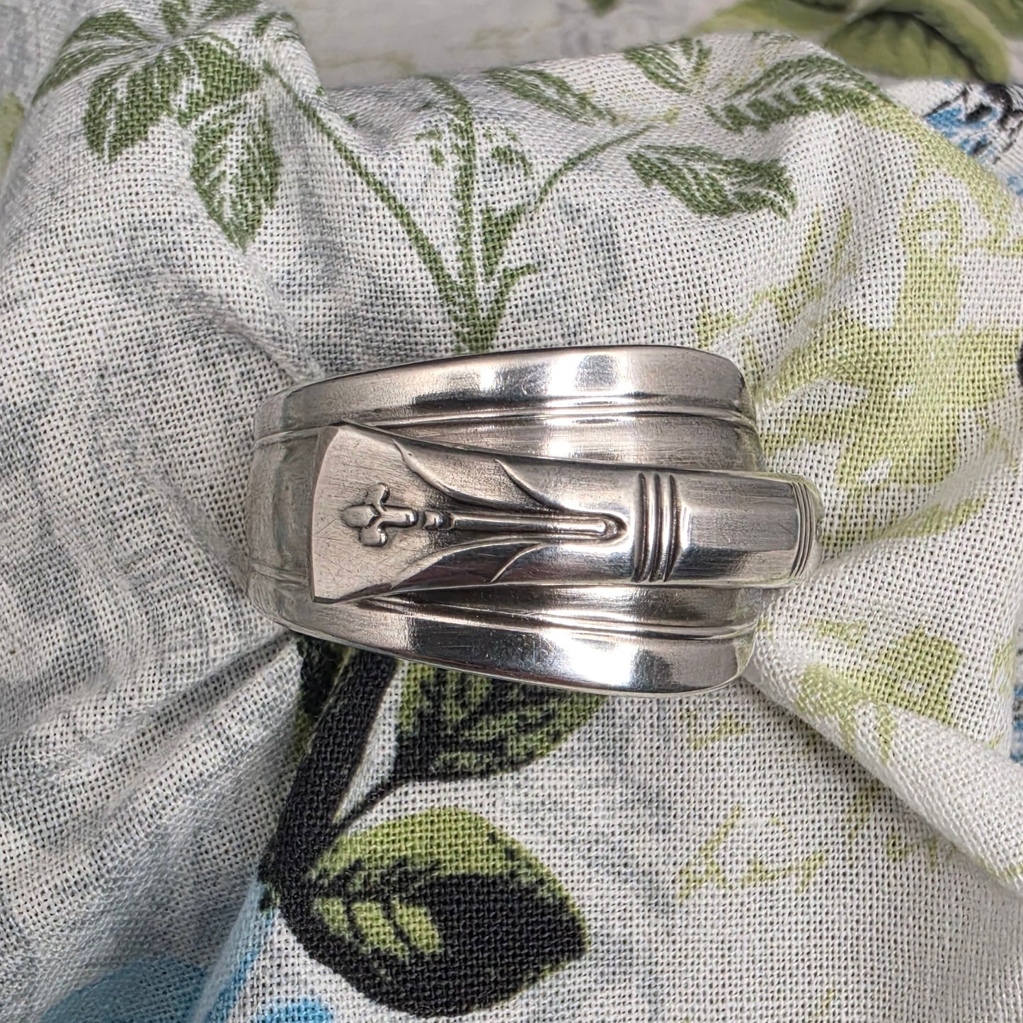 A silver-toned metal ring with a central floral engraving, displayed on a patterned fabric background featuring blue and green floral motifs.