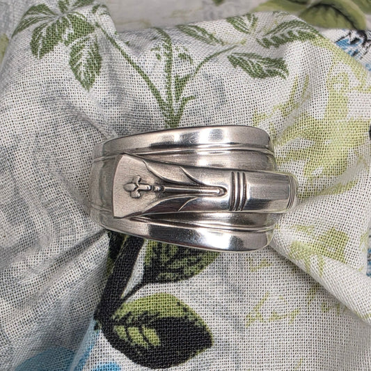 A silver-toned metal ring with a central floral engraving, displayed on a patterned fabric background featuring blue and green floral motifs.