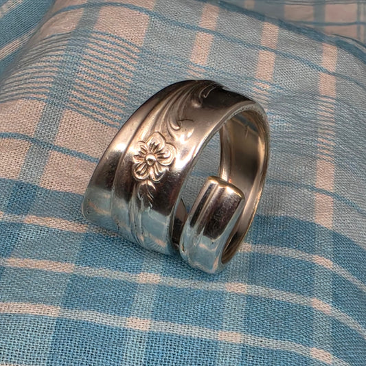 A wide silver-toned metal ring with engraved floral and leaf motifs, photographed on a blue-and-white plaid fabric surface.