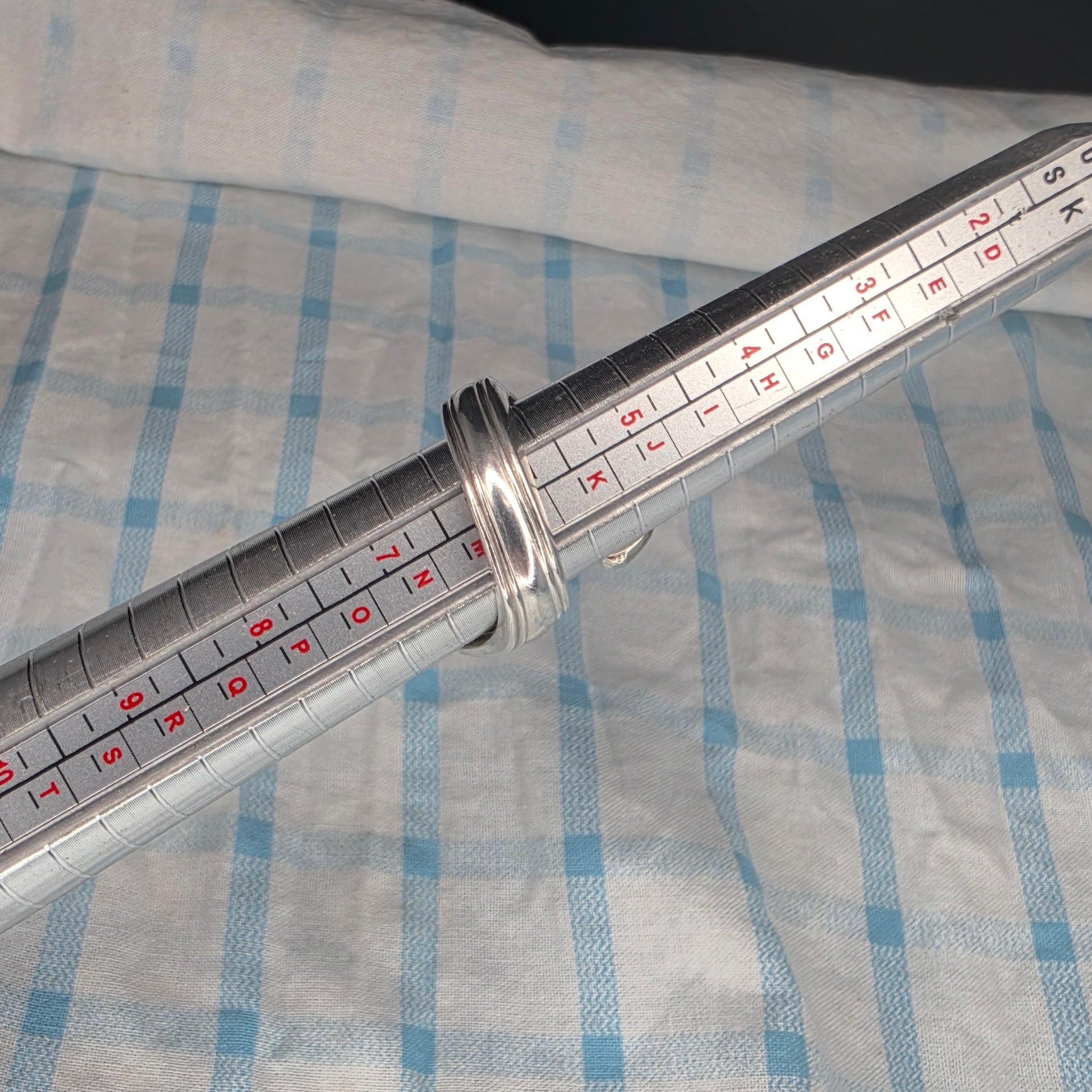A metal ring sizer measuring device with red and black size markings, wrapped with a silver ring to demonstrate use, set against a blue and white checkered fabric background.