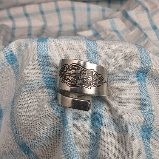 A silver-toned metal cuff ring with an ornate engraved design, resting on a blue and white striped textured fabric.